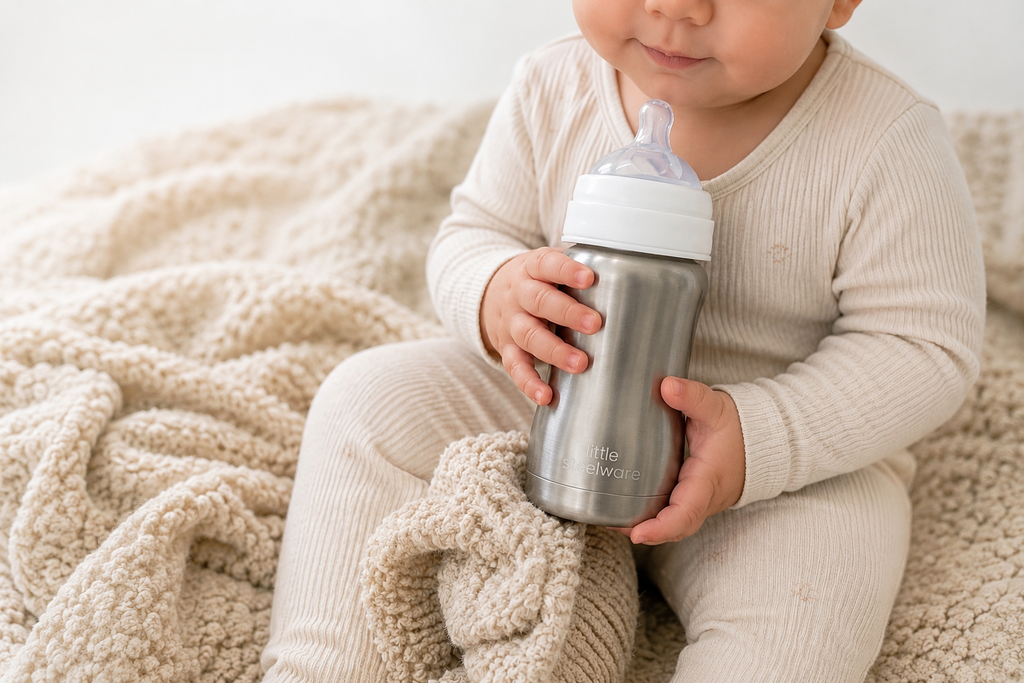 A baby in a cream ribbed onesie holds a stainless steel babu bottle with a white lid and clear nipple, sitting on a soft beige knit blanket.