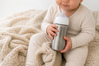 A baby in a cream ribbed onesie holds a stainless steel babu bottle with a white lid and clear nipple, sitting on a soft beige knit blanket.