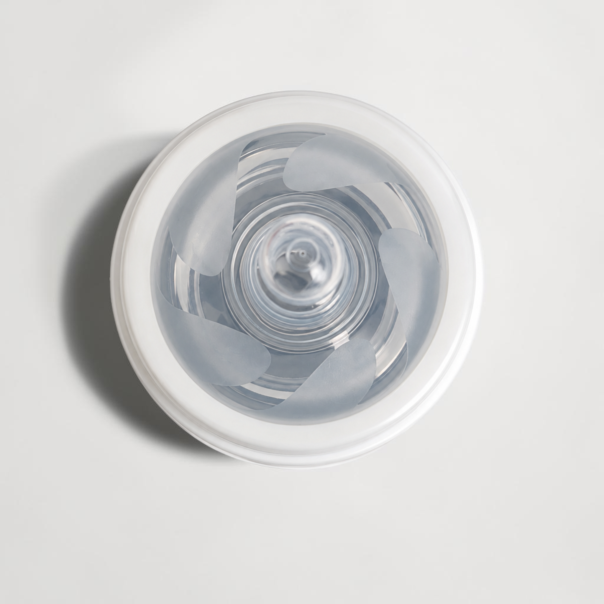 A top-down view of a clear silicone baby bottle nipple inside a white plastic bottle cap, set against a light gray background.