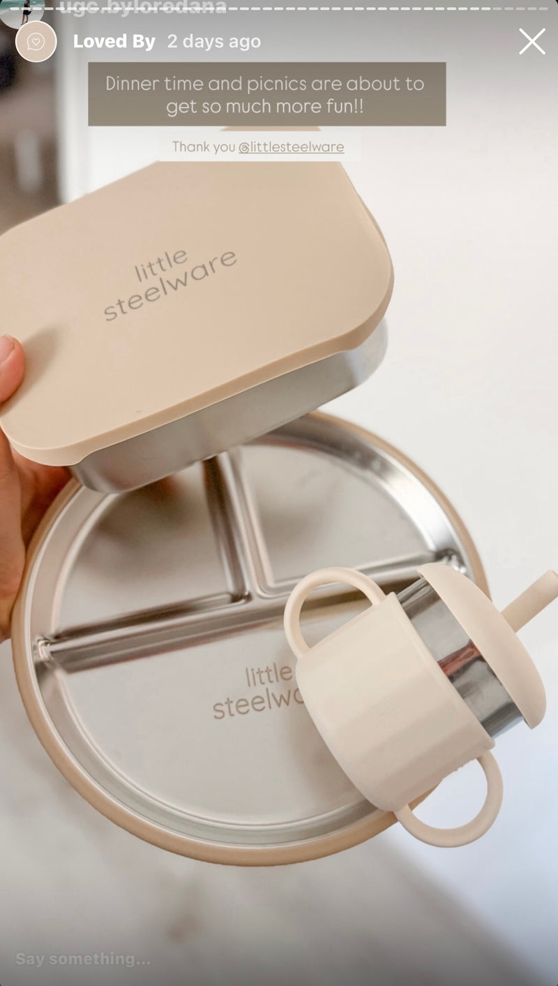 A close-up of a hand holding a beige lid with the brand name 'little steelware' printed on it, above an open round stainless steel food storage container with three compartments and a matching beige cup with a straw attached.