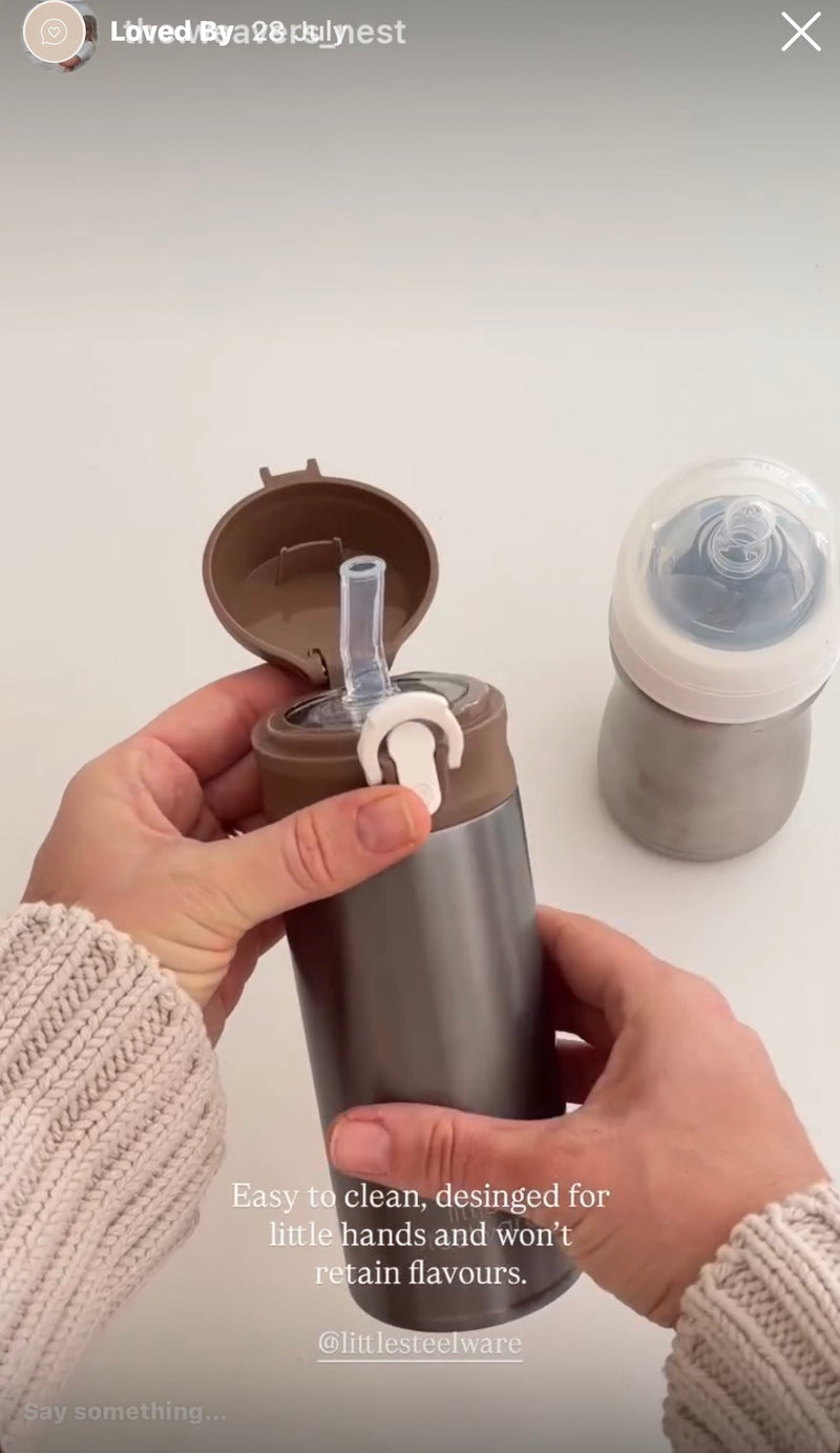 A person's hands holding a stainless steel travel tumbler with a brown lid and clear straw, next to a gray baby bottle on a light gray background.