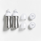 Two stainless steel baby bottles with white caps and clear nipples, shown alongside two clear silicone bottle covers and two clear caps on a clean white background.