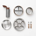 A flat-lay view of a stainless steel lunch set on a white background, including a water bottle, divided plates, a bowl, utensils, and small wooden-handled tools. The brand name 'little stonewall' is visible on the plate.