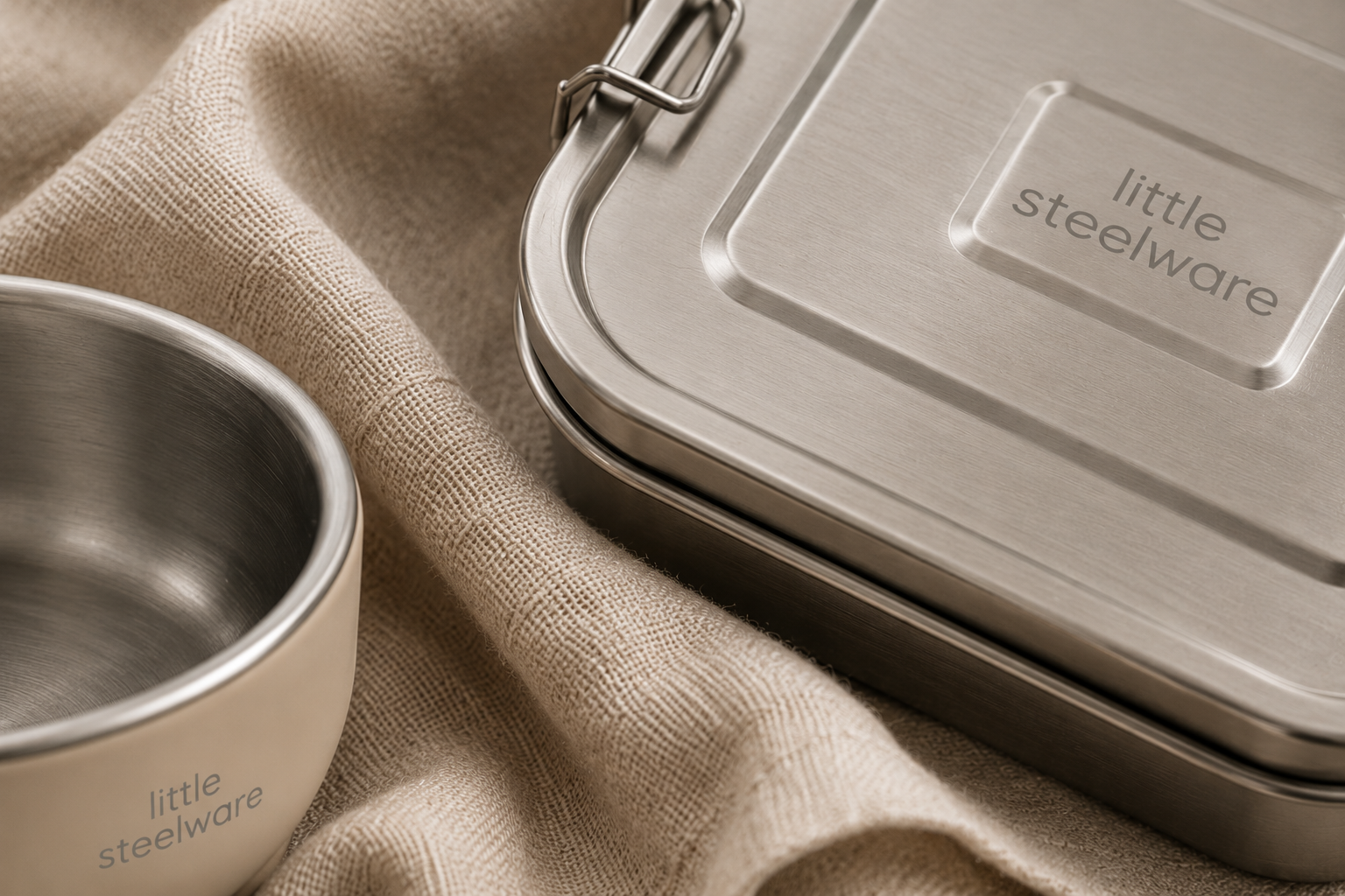 A stainless steel lunch box with a brushed metal lid and a matching beige container, both branded 'little steelware', arranged on a textured beige fabric background.