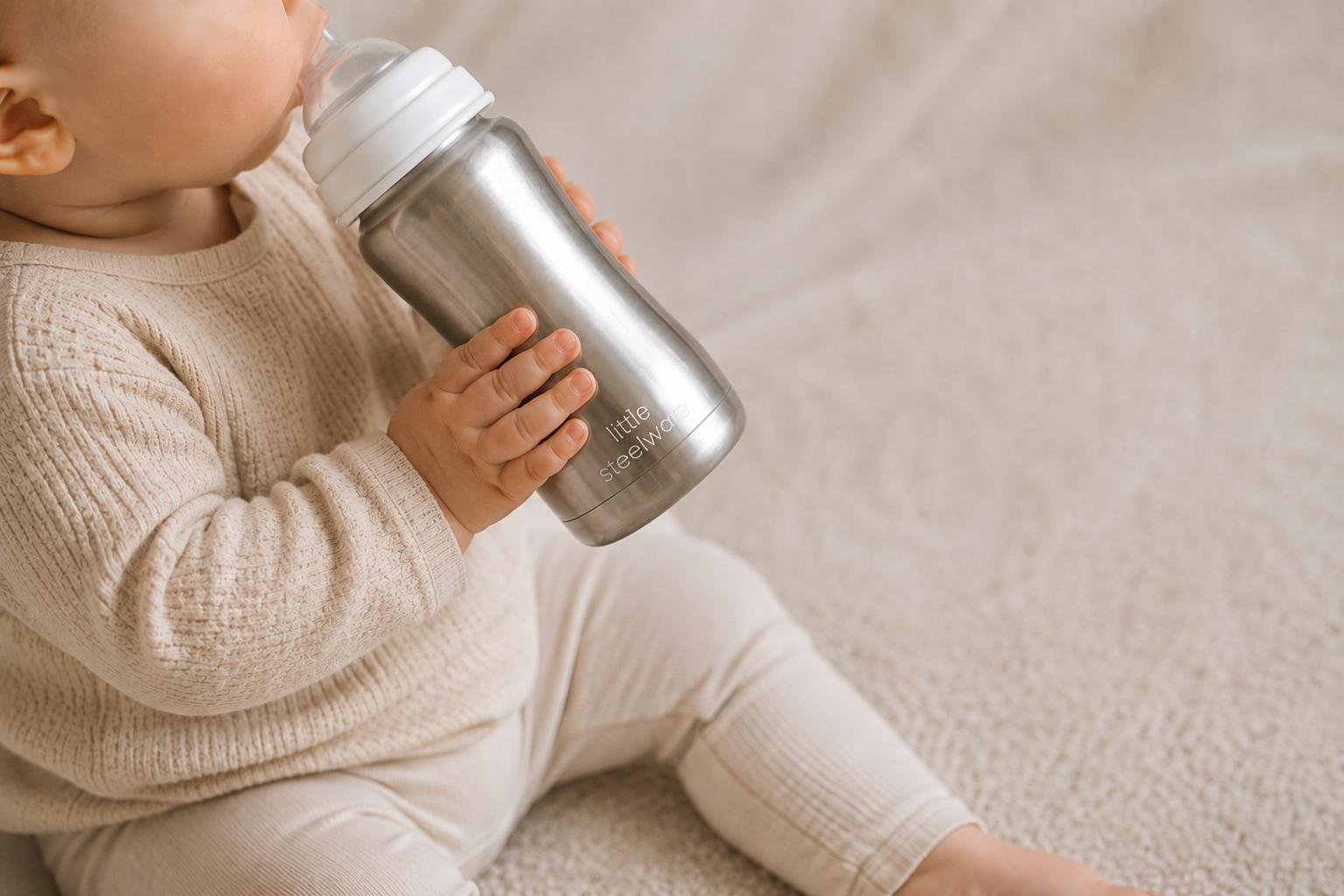 A baby in a cream knit outfit drinking from a brushed stainless steel bottle with a white lid, sitting on a soft beige blanket.
